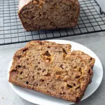 A slice of butterscotch banana bread on a plate with the rest of the loaf on a wire rack behind it.