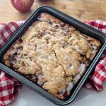 A pan of Apple cinnamon cake on a red and white checkered towel with an apple barely visible behind it.