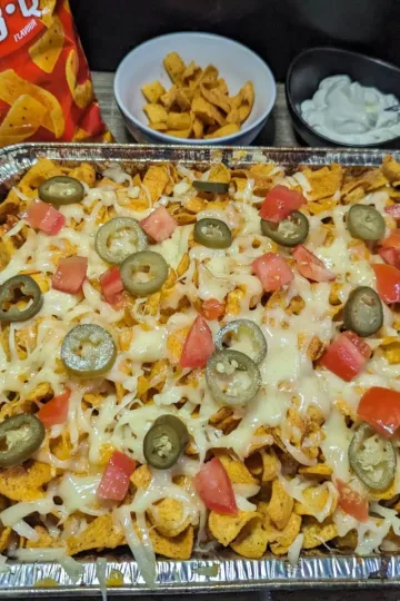 Pan of Fritos Chicken Casserole topped with pickled jalapenos and diced tomatoes. A bag of BBQ Fritos and a bowl of sour cream in the background.