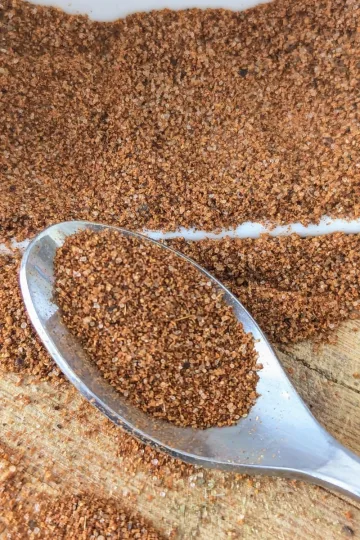 A spoonful of all-purpose seasoning on a board with a white bowl of seasoning spilled behind the spoon.