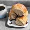 A stack of raisin buns on a white plate with a cup of coffee to the left of the plate.