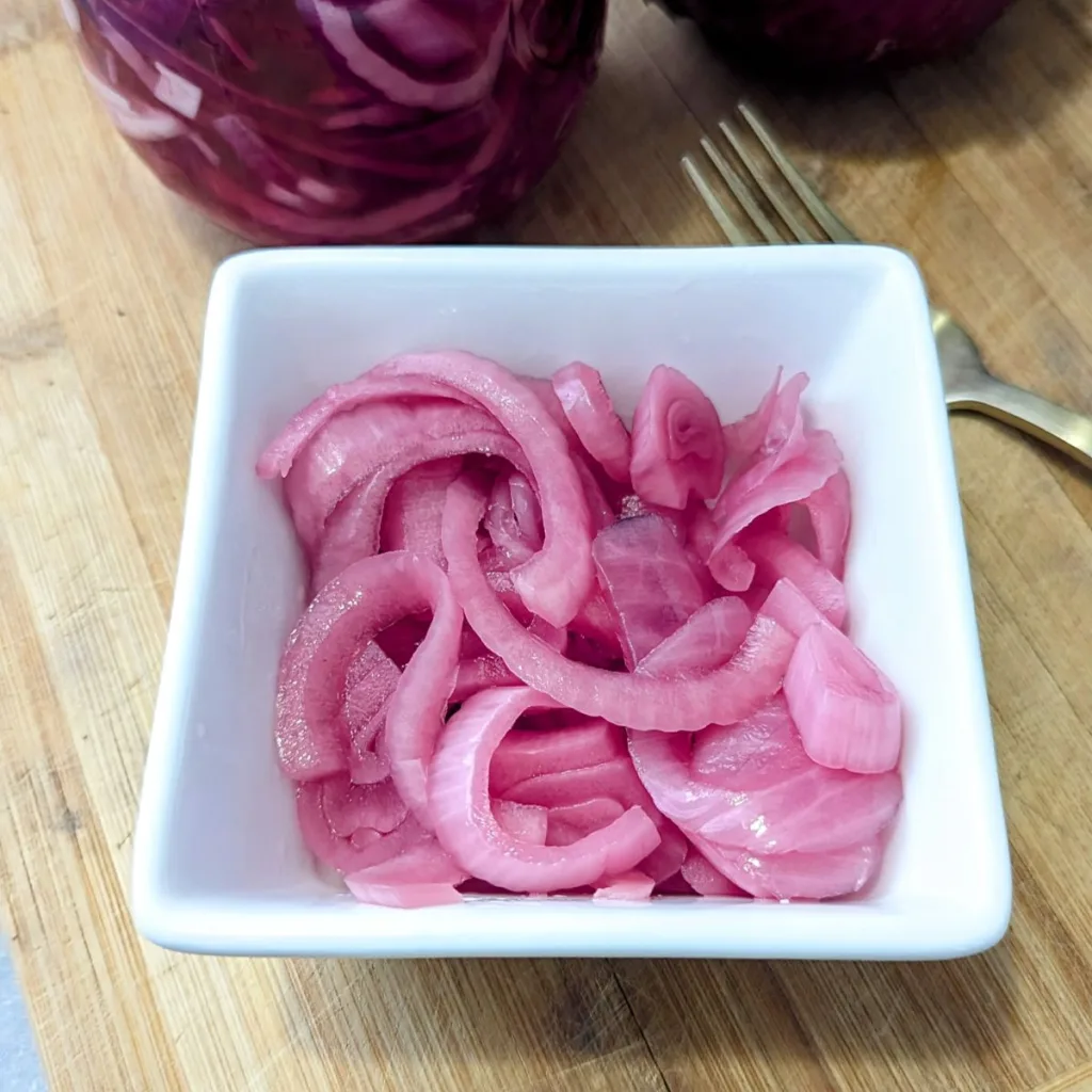 A square white bowl filled with Quick and Easy Pickled Red Onions with the bottom of the jar filled with onions showing behind it.