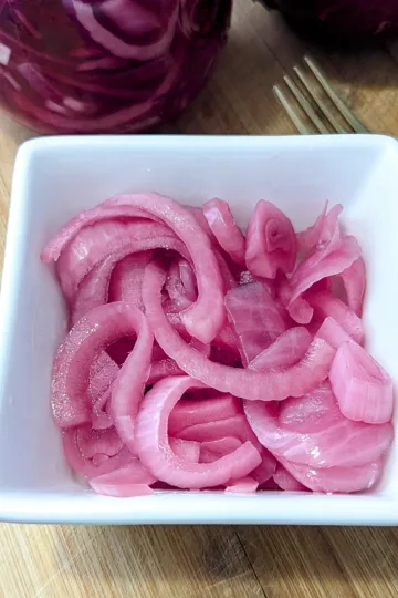 A square white bowl filled with pickled red onions with the bottom of the jar filled with onions showing behind it.