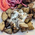 Roasted potatoes with garlic sauce on a plate with tomato slices and pickled red onions.