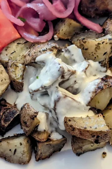 Roasted potatoes with garlic sauce on a plate with tomato slices and pickled red onions.