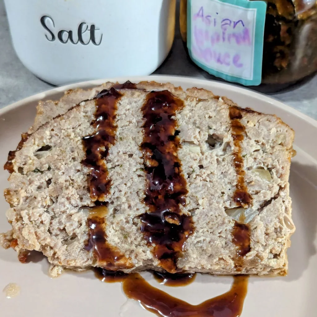 A slice of Glazed Asian-Inspired Pork Meatloaf on a plate with Asian style sauce poured over it.
