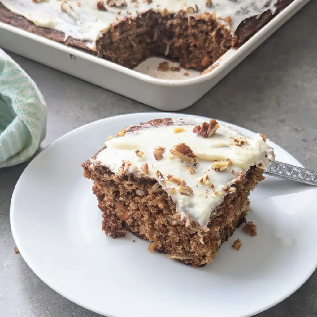 A piece of Carrot Snack Cake on a small white plate with only the handle of a fork showing behind it. The pan of cake is behind the plate with one piece cut out of it.