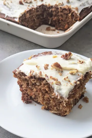 A piece of Carrot Snack Cake on a small white plate with only the handle of a fork showing behind it. The pan of cake is behind the plate with one piece cut out of it.