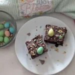 A white plate with two mini egg brownies on it. A bowl of mini eggs is off to the left side and a mint green towel with the words just bloom is at the top of the picture.