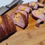 Bacon wrapped stuffed pork tenderloin sliced on a wood cutting board.