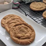 Three sugar and spice cookies on a white plate with a small white bowl of cinnamon sugar and a wire rack of cookies in the background.