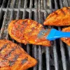 BBQ pineapple chicken breasts on a grill with a blue basting brush adding sauce to the chicken.