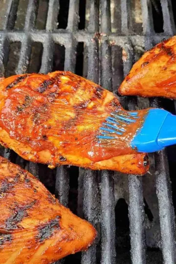 BBQ pineapple chicken breasts on a grill with a blue basting brush adding sauce to the chicken.