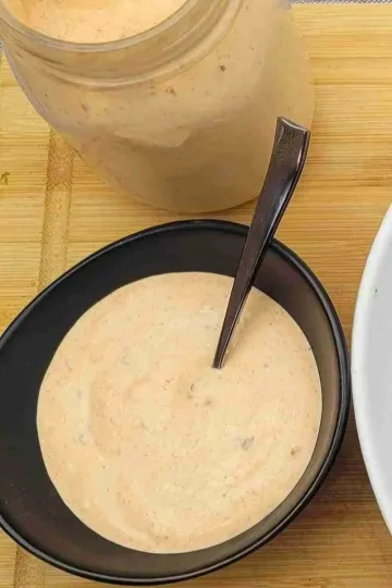 A black bowl filled with homemade chipotle sauce. There is a spoon in the bowl. A jar of the sauce is at the top of the picture and a rim of a white plate is on the right of the bowl with sweet potato fries barely visible.