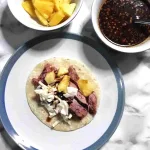 A pork belly taco on a plate with a bowl of fresh pineapple and a bowl of sauce.