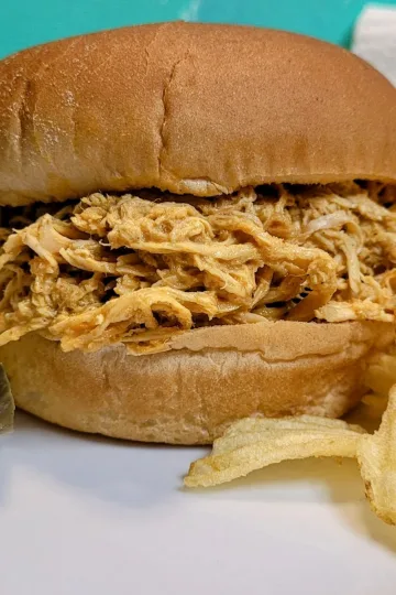 Slow cooker Buffalo chicken on a bun with pickles on the left side and potato chips on the right side.