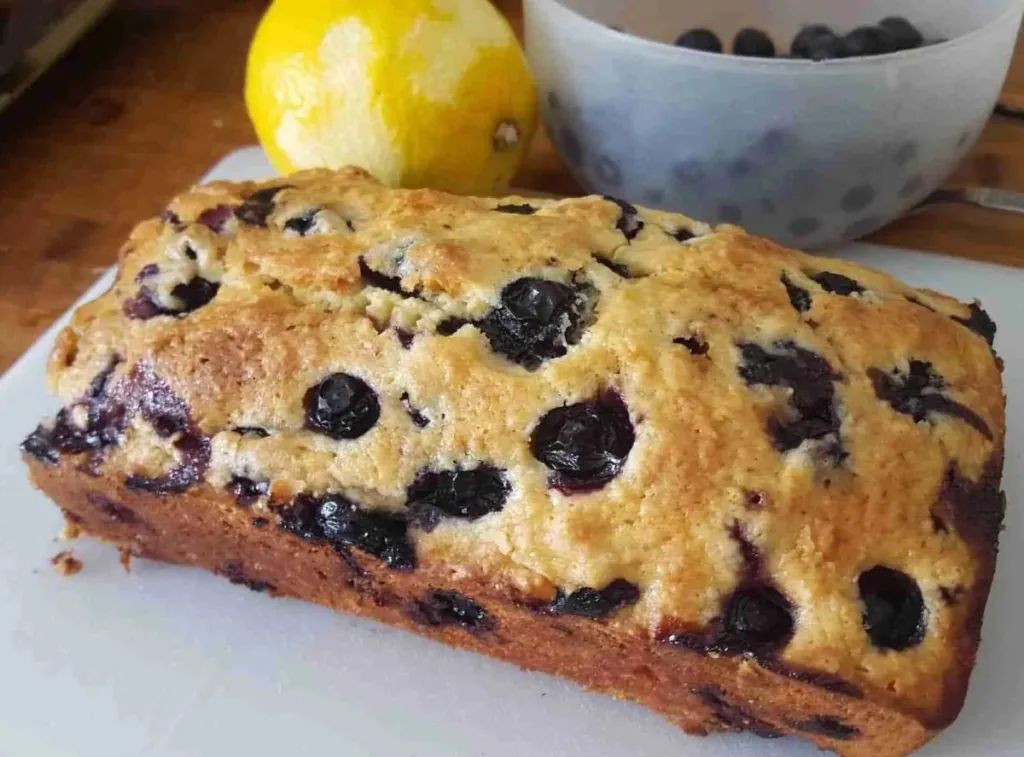 Blueberry lemon loaf on a white board with a lemon and bowl of blueberries behind it.