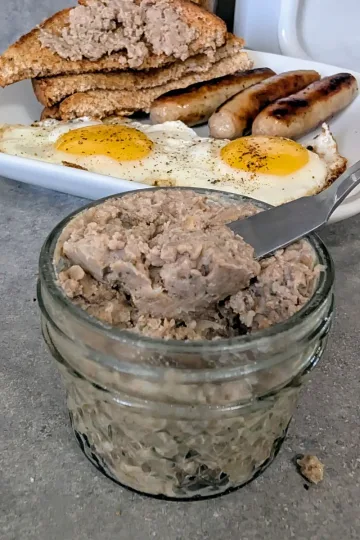 A small jar of French Canadian Cretons with a knife in the jar. There is a plate of fried eggs, sausages and toast with cretons in the background.