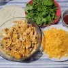 A bowl of easy shredded chicken with a plate of grated cheddar cheese, a bowl of sauce, a plate of lettuce and a pile of tortillas on a white and blue striped cloth.
