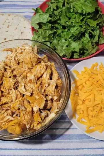 A bowl of easy shredded chicken with a plate of grated cheddar cheese, a bowl of sauce, a plate of lettuce and a pile of tortillas on a white and blue striped cloth.
