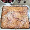 A pan of cherry butter cake sprinkled with powdered sugar on the top. There is a bowl of fresh cherries just visible in the top left and three cherries beside it.