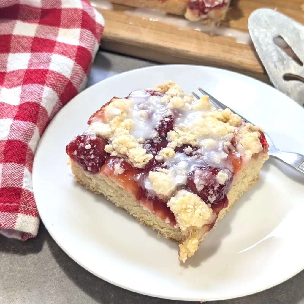 A piece of cherry pie squares on a small white round plate with a fork behind the square. There is a red and white checkered towel to the left, and the edge of a pie server to the right.
