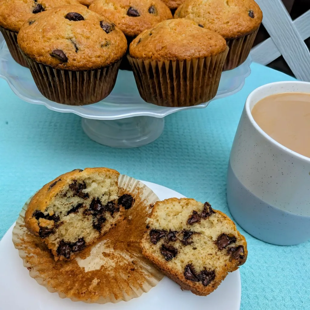 A chocolate chip muffin broken open on a plate, in front of a pedestal plate filled with more muffins and a cup of coffee off to the right.