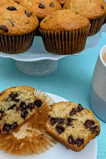 A chocolate chip muffin broken open on a plate, in front of a pedestal plate filled with more muffins and a cup of coffee off to the right.