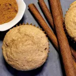 A low sugar cinnamon muffin with cinnamon sticks to the right and a small white bowl of ground cinnamon in the top left corner.
