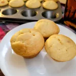 Three pancake muffins on a plate with the remaining muffins in a pan behind.