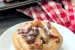 One bacon apple cinnamon rolls on a plate with a pan of rolls behind it on a red and white checkered cloth.