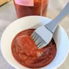 A bowl of Bourbon BBQ sauce with a basting brush in it and a jar behind the bowl.