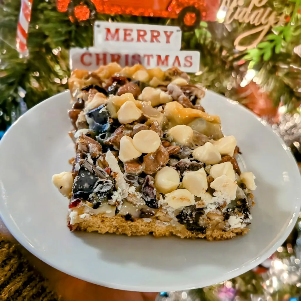 A piece of Layered Christmas Bars on a white plate with a close up of a Christmas tree in behind the plate. There is an ornament that says Merry Christmas in red writing on a white background.