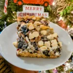 A piece of Layered Christmas Bars on a white plate with a close up of a Christmas tree in behind the plate. There is an ornament that says Merry Christmas in red writing on a white background.