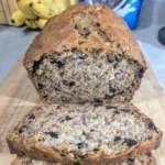 A loaf of Oreo Banana Bread sliced on a wood cutting board. There is a bunch of bananas behind the loaf on the left and the bottom of an Oreo cookie bag is barely visible in the top right beside the bananas.