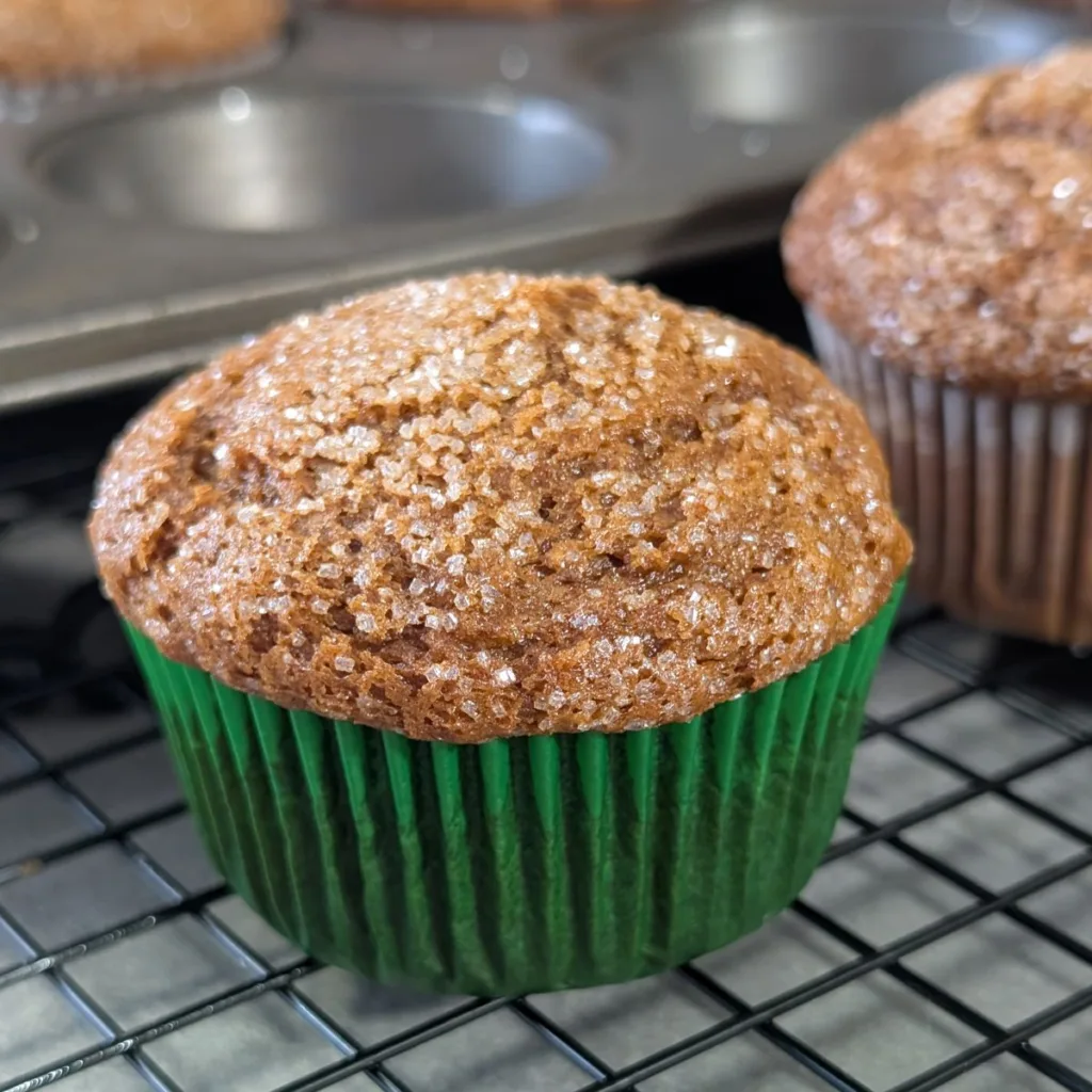 A gingerbread muffin in a green muffin paper, resting on a wire rack with the muffin pan behind barely visible missing two muffins.