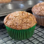 A gingerbread muffin in a green muffin paper, resting on a wire rack with the muffin pan behind barely visible missing two muffins.