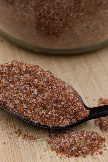 A spoonful of all-purpose BBQ seasoning on a wooden cutting board and the bottom of the jar of seasoning behind the spoon.