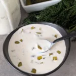 A bowl of Dill Pickle Sauce with a spoon. There is fresh dill and a jar of sauce in the back of the bowl.