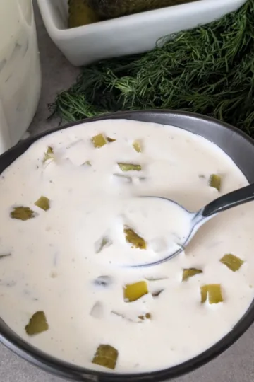 A bowl of Dill Pickle Sauce with a spoon. There is fresh dill and a jar of sauce in the back of the bowl.