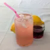 A glass of Homemade Pink Lemonade with ice and a pink straw. There are two lemons and a small jar of grenadine behind the glass.