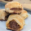 Four homemade sausage rolls on a plate. They are piled as three on the bottom and one on the top of the others.