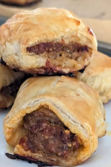 Four homemade sausage rolls on a plate. They are piled as three on the bottom and one on the top of the others.