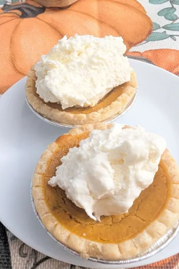 Two mini pumpkin pies with whipped cream topping on a white plate set on a fall pumpkin tablecloth.