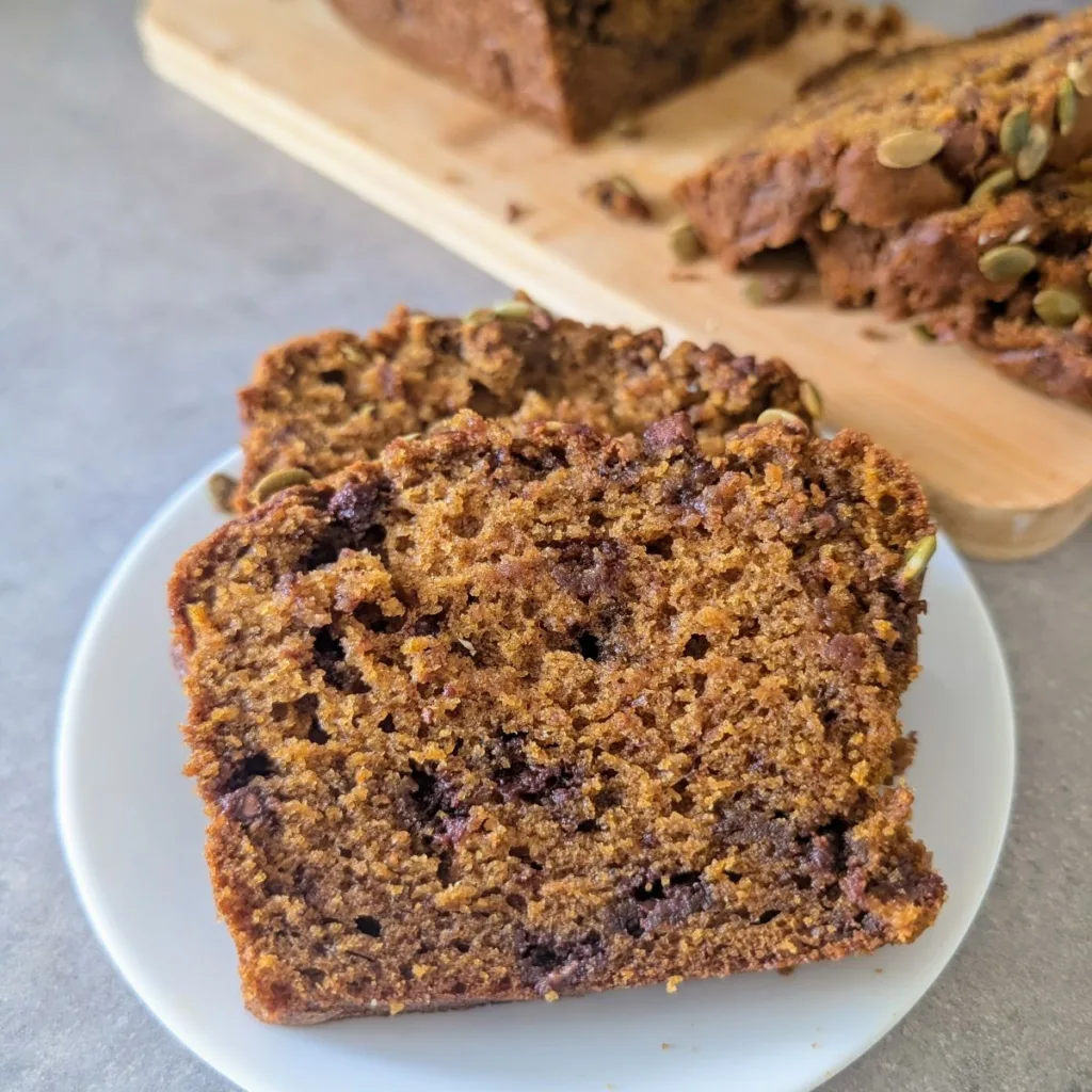 Two slices of Pumpkin Chocolate Chip Bread on a plate with the rest of the loaf in the background on a cutting board.
